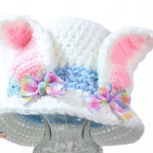 May include: White crocheted bunny hat with pink ears and a blue and white striped band. The hat is decorated with two pink and white gingham bows.