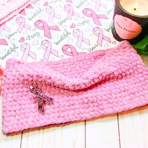 May include: A pink, textured ear warmer with a sparkling pink ribbon pin. The ear warmer is displayed on a wooden surface with a floral arrangement, a candle, and a patterned paper with pink ribbons and the words "Brave, Beautiful, Strong".