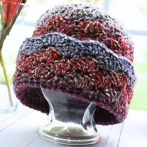 May include: A crocheted hat featuring a variegated yarn pattern in shades of deep red, charcoal gray, and metallic gold. The hat has a wide, folded brim with a textured stitch pattern and is displayed on a clear, glass-like head form.
