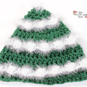 Op de afbeelding: Groene gehaakte kerstboomhoed met witte gehaakte rand en wit glinsterend garen. De hoed is gevormd als een kerstboom en heeft drie lagen rand.
