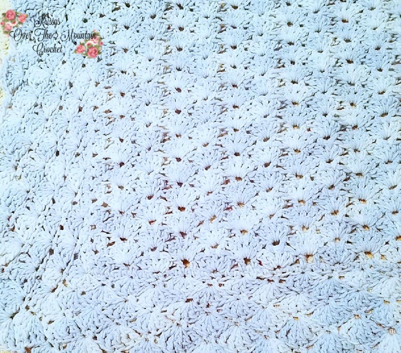 May include: A close-up of a light blue crocheted blanket with a repeating pattern of small flowers. The blanket is made with a soft, fluffy yarn.