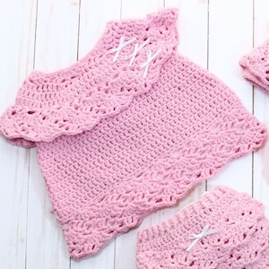 May include: A pink crocheted baby outfit with a ruffled top and a matching diaper cover. The top has white satin bows on the neckline.
