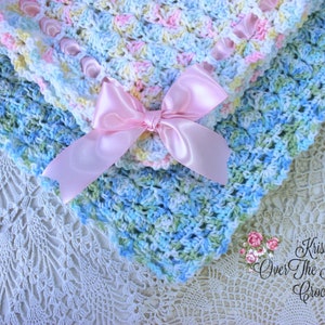 May include: A crocheted baby blanket in shades of blue, green, and pink with a pink satin ribbon tied in a bow. The blanket is on a white crocheted doily. The text "Krissys Over The Mountain Crochet" is visible in the lower right corner of the image.
