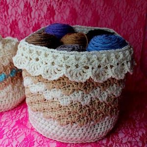 May include: A handmade crochet basket with a scalloped edge, filled with various colors of yarn. The basket features alternating bands of white, beige, and brown. The yarn colors include blue, brown, and purple.