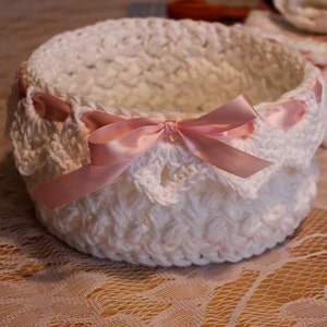 May include: White crocheted basket with a pink satin ribbon. The basket has a scalloped edge and is perfect for holding small items.