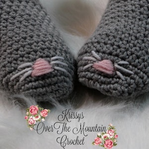 May include: A pair of gray crocheted slippers with pink and white embroidered cat faces. The slippers are on a white fuzzy surface.
