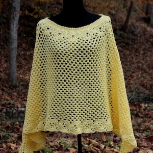 May include: A yellow crochet poncho with a delicate openwork pattern. The poncho is displayed on a black mannequin against a backdrop of fallen leaves.
