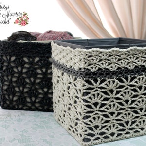 May include: Two square storage baskets with crocheted covers. The basket on the left is black and the basket on the right is beige with a black crocheted band around the middle.