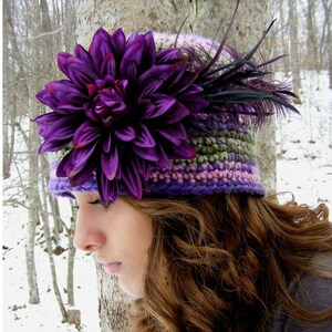 Puede incluir: Un gorro de punto de color morado y verde con una gran flor morada y plumas.