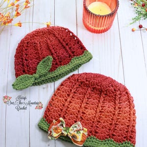 May include: Two crocheted hats with a ribbed texture in shades of red and orange. One hat features a green crocheted leaf accent, while the other has a fabric bow with a pumpkin and sunflower print. Both hats have a green crocheted brim.