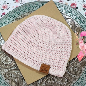 K&ouml;nnte beinhalten: Rosa gestrickte M&uuml;tze mit einem braunen Lederanh&auml;nger, auf dem "stitched with love" steht. Die M&uuml;tze liegt auf einer braunen Oberfl&auml;che mit einem rosa Band mit Blumen im Hintergrund.