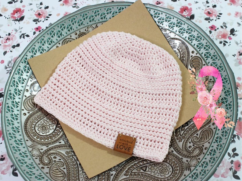 K&ouml;nnte beinhalten: Rosa gestrickte M&uuml;tze mit einem braunen Lederanh&auml;nger, auf dem "stitched with love" steht. Die M&uuml;tze liegt auf einer braunen Oberfl&auml;che mit einem rosa Band mit Blumen im Hintergrund.