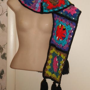 May include: A colorful, handmade crochet scarf with a square pattern. The scarf features a variety of colors including blue, red, green, and purple, with black borders and tassels. The scarf is displayed on a mannequin.