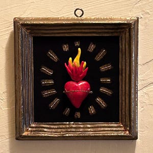 May include: A square wall hanging featuring a red heart with yellow flames and a wire crown of thorns, centered on a black background. The heart is surrounded by twelve small, dark, textured rectangular pieces arranged in a circle, all encased in a rustic, dark metallic frame with a top hanging loop.