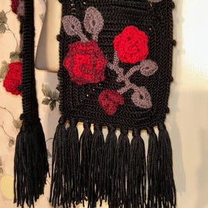 May include: Black crochet scarf with red and burgundy rose appliques and gray leaves. The scarf has a fringe of black tassels at the bottom. The scarf is displayed on a mannequin.