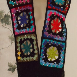 May include: A colorful crocheted scarf with a fringe. The scarf is made of black yarn with squares of different colors, including red, blue, green, yellow, and purple. Each square has a star-shaped design in the center.