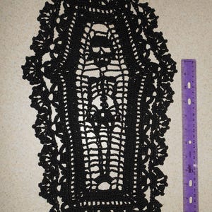 May include: Black crochet coffin-shaped doily with intricate openwork design and scalloped edges. A purple ruler is placed next to the doily for scale. The doily is approximately 10 inches long.