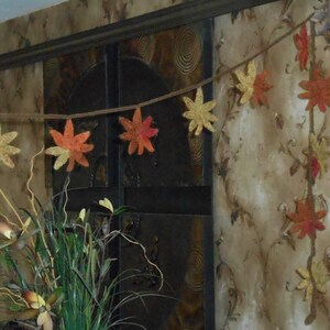 May include: A garland of crocheted fall leaves in shades of orange, yellow, and brown hangs against a textured wall. The leaves are strung on a twine cord and are arranged in a staggered pattern.