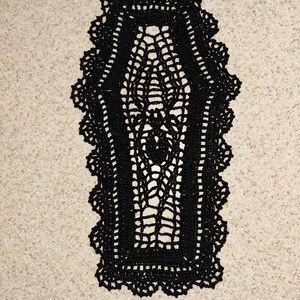 May include: A black, crocheted coffin-shaped doily with a delicate, open-weave design. The doily features a scalloped edge and a spiderweb-like pattern in the center, perfect for Halloween or gothic decor.