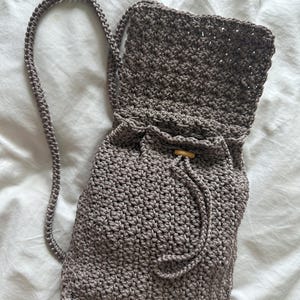 Peut inclure: Un sac bandoulière en crochet de couleur taupe avec un motif de points texturés, une longue bandoulière tressée et une fermeture à cordon sécurisée par une petite perle en bois. Le sac présente une base rectangulaire et un rabat supérieur.