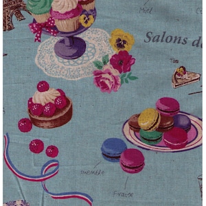 May include: A blue and white fabric with a French theme. The fabric features images of cupcakes, macarons, a slice of cake, and a Eiffel Tower. The fabric also has the words "Salons de", "Miel", "Fraises", "Menthe", and "Bon", written in French.