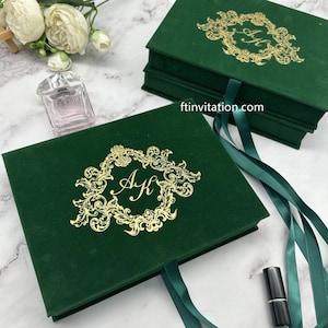 May include: Emerald green velvet invitation boxes with gold foil monogram and decorative border. The boxes have a ribbon closure and are stacked. A bottle of perfume and lipstick are also in the image.