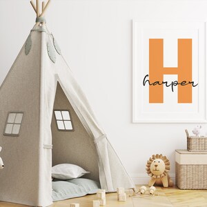 Op de afbeelding: Een kinderkamerscène met een beige tipi-tent, een oranje ingelijste print met de letter "H" en de naam "harper", en een knuffel leeuw. Een wit kangoeroe speelgoed en houten blokken zijn ook zichtbaar.