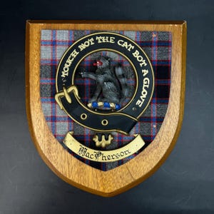 May include: Wooden plaque with a tartan background and a black emblem featuring a cat figure. The emblem has the text "TOUCH NOT THE CAT BOT A GLOVE" and "MacPherson" on a banner.