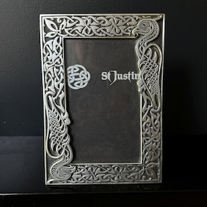 May include: Silver metal picture frame with a detailed Celtic knotwork design. The rectangular frame has a dark interior and the text "St Justin". The border features interwoven patterns and stylized animal figures, showcasing traditional Celtic artistry.