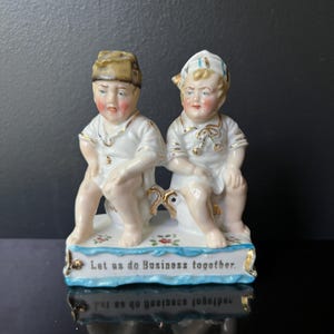 May include: A vintage porcelain figurine of two cherubic figures in white outfits with gold trim, seated together. The base is decorated with floral accents and the text "Let us do Business together." The figurine has a glossy finish and is set against a dark background.