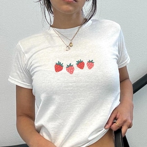 May include: White t-shirt featuring a row of five embroidered strawberries. Each strawberry has a red body and green leaves. The shirt is short-sleeved and made of a soft material.
