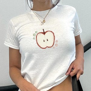 May include: White t-shirt with a hand-drawn apple design. The apple is red with a white interior and two black seeds. The design includes two green and two pink button-like shapes. The shirt has short sleeves.