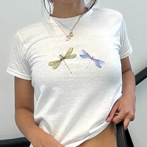 May include: White t-shirt with two dragonfly designs. One dragonfly is green and yellow, the other is blue and purple. The t-shirt has a crew neck and is made of a soft material.