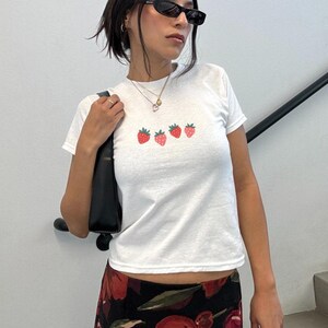 May include: White t-shirt with a crew neck featuring four embroidered strawberries in a row. The strawberries are red with green stems. The shirt is paired with a black purse and a floral print skirt.