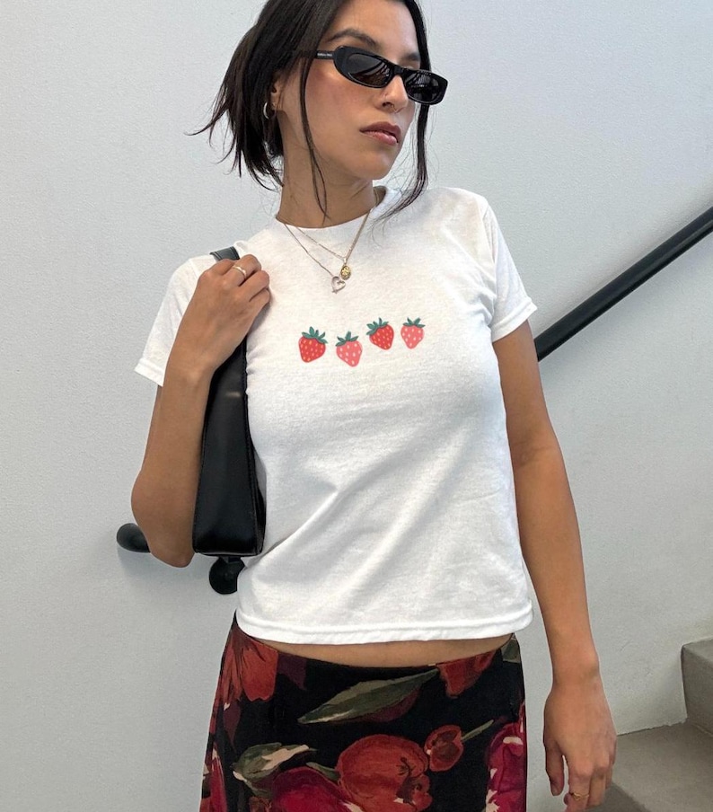 May include: White t-shirt with a crew neck featuring four embroidered strawberries in a row. The strawberries are red with green stems. The shirt is paired with a black purse and a floral print skirt.