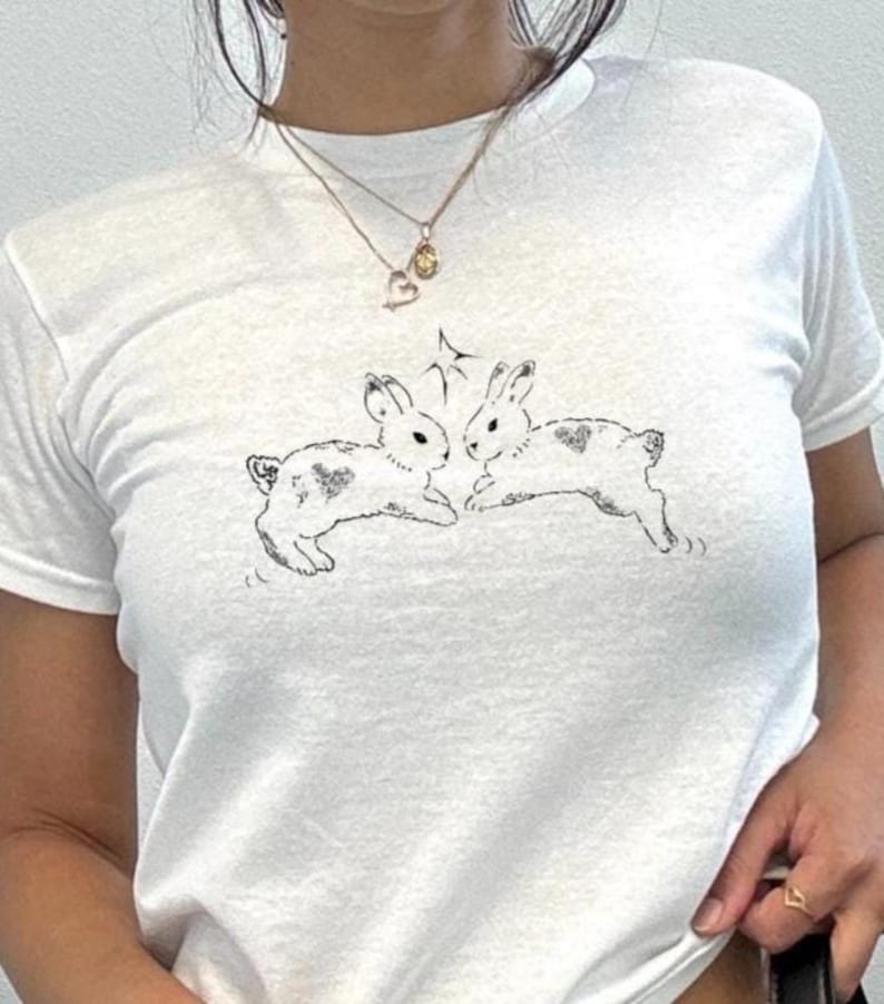 Puede incluir: Camiseta blanca con una ilustraci&oacute;n en blanco y negro de dos conejos. Cada conejo tiene una marca en forma de coraz&oacute;n. Un dise&ntilde;o de estrella est&aacute; encima de los conejos, que est&aacute;n en una pose juguetona.