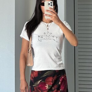 Puede incluir: Camiseta blanca de manga corta con un gr&aacute;fico de conejos en blanco y negro. La persona lleva una falda floral con flores rojas y negras, un peque&ntilde;o bolso beige y gafas de sol. La persona sostiene un tel&eacute;fono.