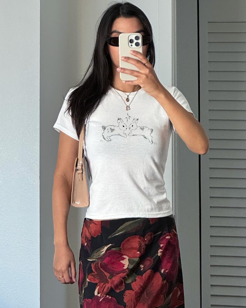 Puede incluir: Camiseta blanca de manga corta con un gr&aacute;fico de conejos en blanco y negro. La persona lleva una falda floral con flores rojas y negras, un peque&ntilde;o bolso beige y gafas de sol. La persona sostiene un tel&eacute;fono.