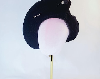 May include: A black velvet hat with decorative pins, displayed on a pink mannequin head atop a gold-colored stand. The hat has a curved, asymmetrical shape and is adorned with two silver pins and a small, beaded embellishment.