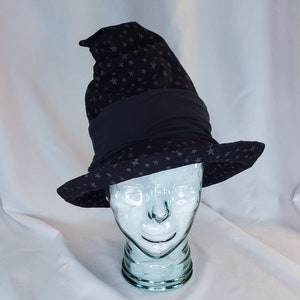 May include: A black witch hat with a star pattern. The hat has a wide brim and a tall, pointed crown. A black band encircles the base of the crown. The hat is displayed on a clear glass head.