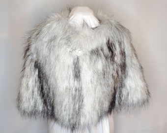 Peut inclure: Une cape en fausse fourrure dans les tons de gris et de blanc, avec un motif sombre. La cape est fermée par un fermoir décoratif au niveau de l'encolure. Le vêtement est présenté sur un mannequin blanc.