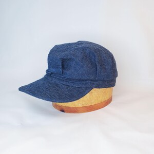 Peut inclure: Casquette de style gavroche en denim bleu foncé avec une petite visière. Le chapeau est fait d'un tissu texturé et repose sur une forme en bois. La casquette a une couronne arrondie et une petite visière incurvée.