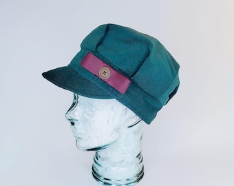 May include: A teal corduroy hat with a short brim and a decorative burgundy band. The band is accented with a wooden button. The hat is displayed on a glass head form against a white background.