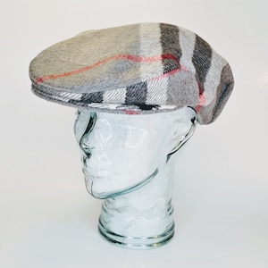May include: A grey flat cap featuring a plaid design in grey, black, and red. The cap is displayed on a clear glass head. The hat has a classic design, suitable for various occasions.