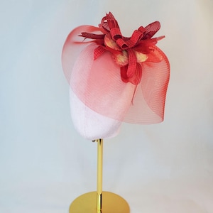May include: A red fascinator hat with a mesh veil and floral embellishments. The hat is displayed on a white mannequin head on a gold-coloured stand. The veil is a sheer, open weave.