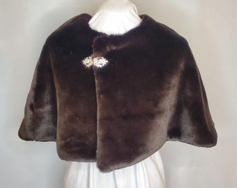 May include: A brown faux fur cape with a decorative clasp. The cape is a dark brown color and has a soft, plush texture. The clasp is gold-toned with clear crystal accents. The cape is designed to be worn over the shoulders.