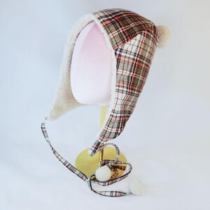May include: A tartan trapper hat with ear flaps and a faux fur lining. The hat features a pattern of red, white, and green squares. The ear flaps are lined with a soft, cream-coloured material. The hat has two cream-coloured pom-poms.