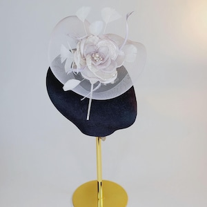 May include: A formal hat on a gold stand. The hat has a black base, a large, pale purple flower with pearl detailing, white feathers, and a white mesh veil. Suitable for special events and occasions.