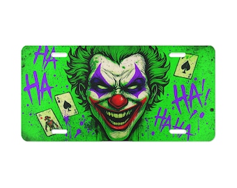 May include: A green license plate featuring a detailed illustration of a clown with green hair, purple accents, and a red nose. The word "HA" is written in purple, along with playing cards. The clown has a sinister grin.