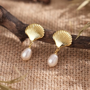 Peut inclure: Une paire de boucles d'oreilles en forme de coquillage dorées, ornées de perles en forme de goutte. Les boucles d'oreilles sont présentées sur une branche en bois, sur un fond texturé de couleur neutre. Un bijou classique.